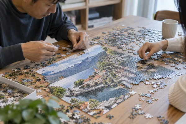adult hands assembling a detailed 1000-piece-style cardboard puzzle at a broad table